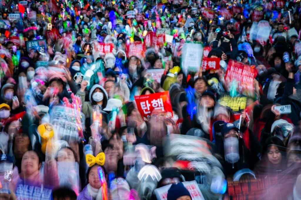 다양한 색의 응원봉과 피켓을 든 시민들을 위에서 찍은 사진. 빨간 피켓에 탄핵이라고 적힌 글씨가 도드라지고, 사람들이 위아래로 뛰고 있어 잔상이 남았다.