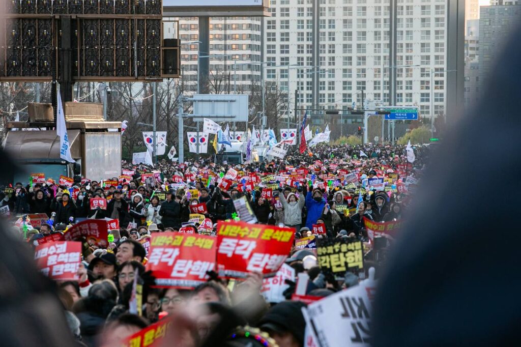 무대 위에서 집회에 참여한 시민들을 찍은 사진. 멀리 아파트와 도로 표지판들이 보이고, 가로등에 태극기가 걸려있다. 가까이에는 시민들이 가득하다. 내란수괴 윤석열 즉각 탄핵이라고 적힌 빨간 피켓을 든 시민들이 도드라진다.