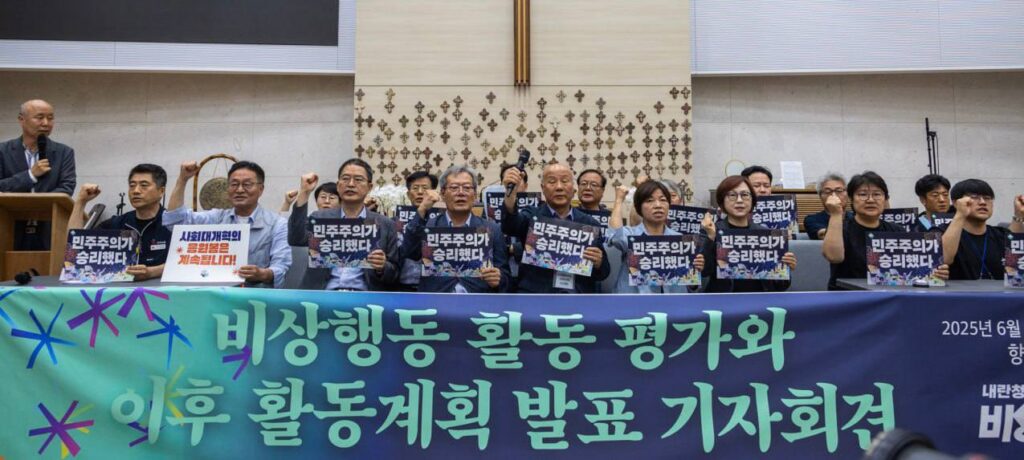 현수막 뒤에 피켓을 들고 앉아 팔을 치켜든 사람들. 비상행동 활동평가와 이후 활동계획 발표 기자회견이라는 글씨가 현수막에 써있다.
