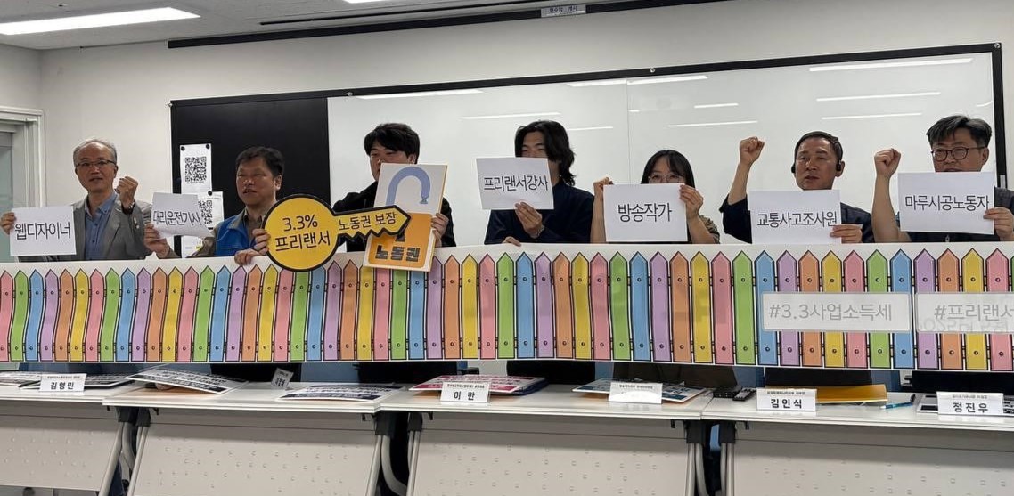 무지개 색깔의 울타리가 그려진 현수막을 든 기자회견 참가자들의 모습. 가운데 참가자는 '노동권'이라고 적힌 자물쇠 모형 판넬과 '3.3% 프리랜서 노동권보장'이 적힌 열쇠 모양의 판넬을 들고 서있다. 다른 참가자들은 프리랜서 직종으로 대표되는 직업군이 적힌 종이를 들고 있다.(웹디자이너, 대리운전기사, 프리랜서 강사, 방송작가, 교통사고조사원, 마루시공노동자)