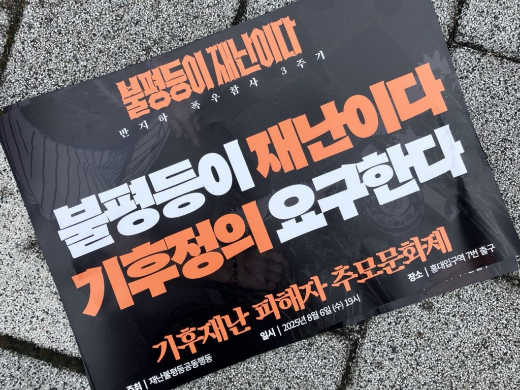 보도블럭에 피켓 한 장이 놓여있다. 검정색 바탕의 피켓 중앙 상단에는 불평등이 재난이다. 반지하 폭우참사 3주기라고 적혀있다. 가운데에는 불평등이 재난이다 기후정의 요구한다라는 글자가 크게 적혀있다. 맨 하단에는 기후재난 피해자 추모문화제 주최 | 재난불평등공동행동 일시 | 2025년 8월 6일(수) 19시 장소 | 홍대입구역 7번 출구라고 적혀있다.