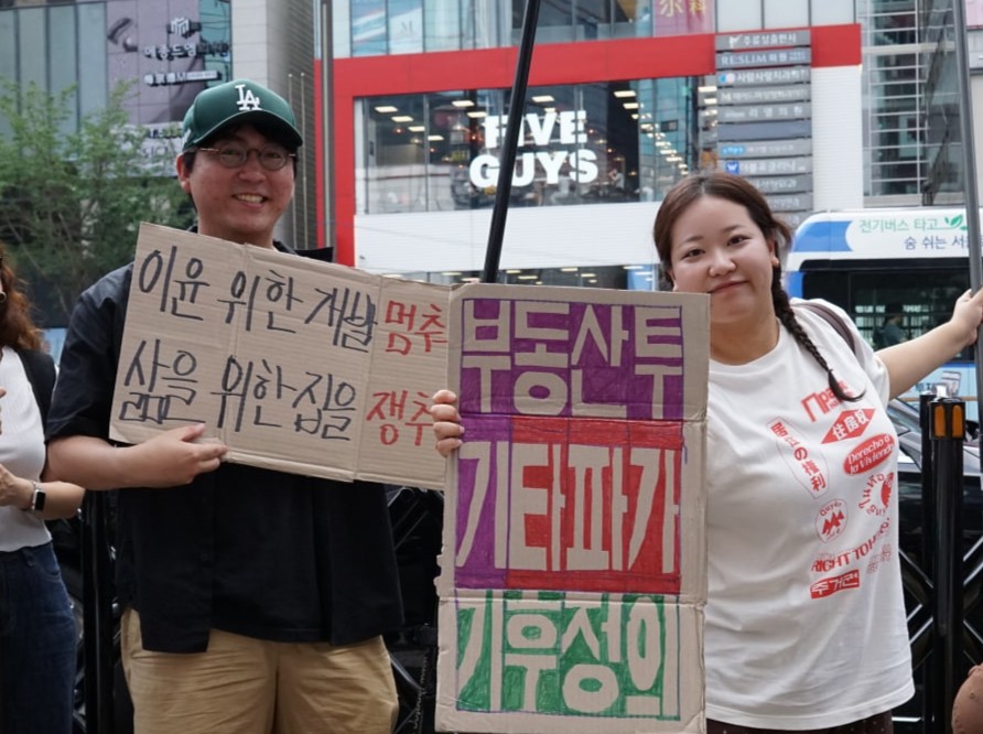 활동가 두 명이 박스로 만든 피켓을 들고 웃고 있다. 왼쪽에 서있는 활동가의 손에는 이윤 위한 개발 멈춰, 삶을 위한 집을 쟁취라고 적혀있고, 오른쪽 활동가의 손에는 부동산 투기 타파가 기후정의라고 적힌 박스 피켓을 들고 있다.